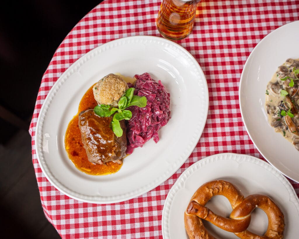 A white plate with braised meat in brown sauce, a bread dumpling, and red cabbage sits on a red and white chequered tablecloth, next to dishes of a soft pretzel and a creamy mushroom dish.