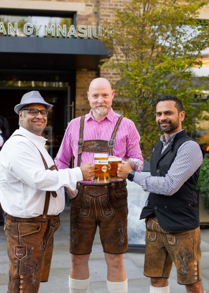 Three men stand outside a building, smiling and holding large beer tankards. Two wear traditional lederhosen outfits, while the third wears a modern waistcoat and shirt. Greenery and a sign reading 