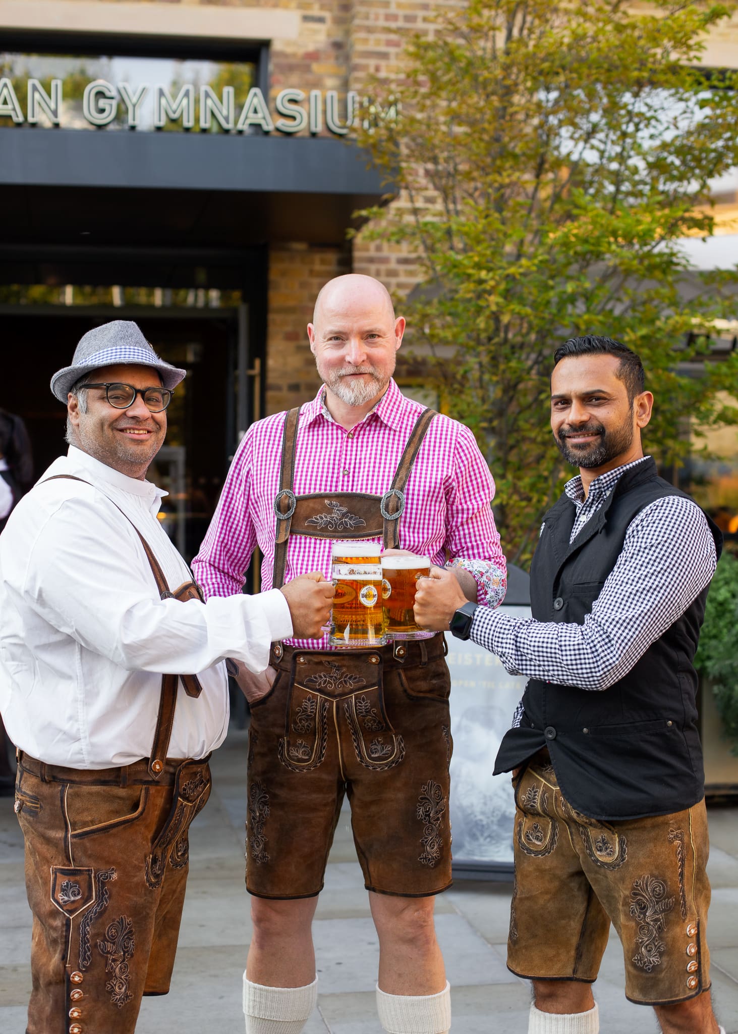 Three men stand outside a building, smiling and holding large beer tankards. Two wear traditional lederhosen outfits, while the third wears a modern waistcoat and shirt. Greenery and a sign reading 