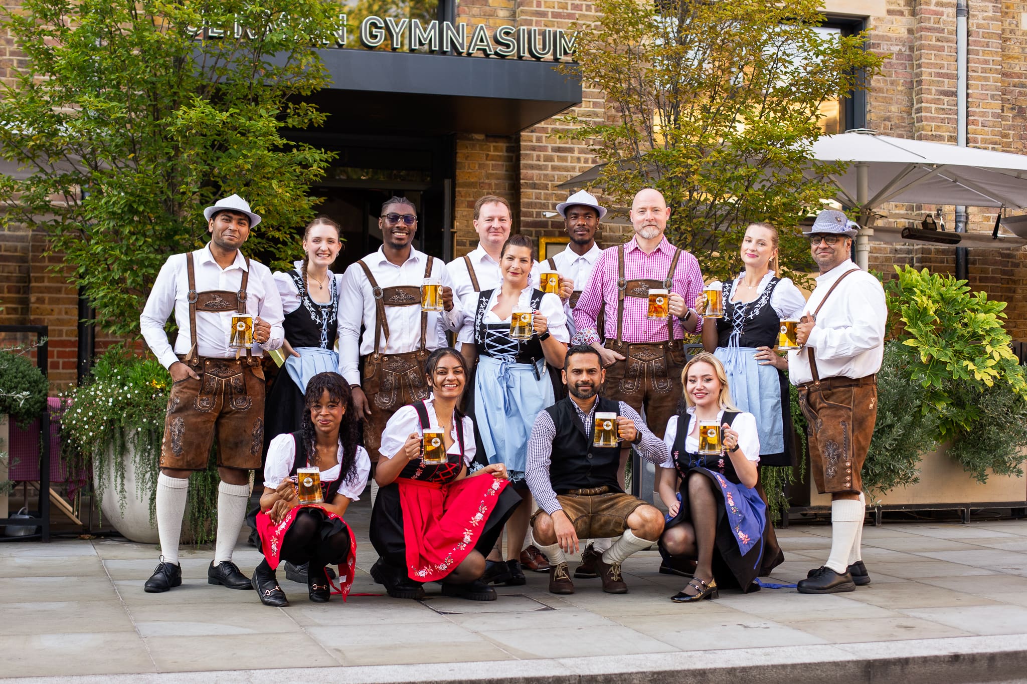 A group of people dressed in traditional Bavarian clothing pose together outdoors, each holding a beer stein and smiling at the camera. They are standing and kneeling on a city pavement in front of a brick building.