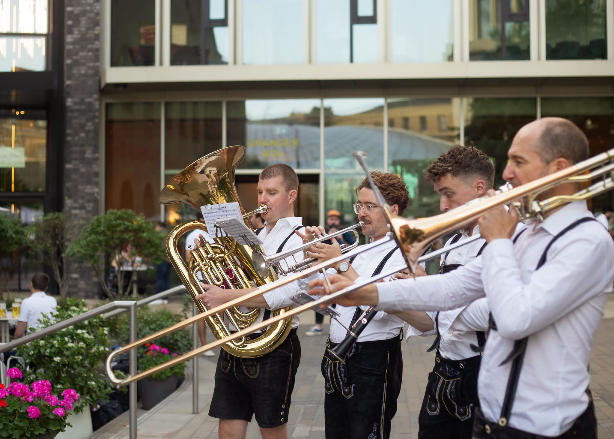 Four musicians in white shirts and braces play brass instruments outdoors, including a tuba and trombones, in front of a modern building with large windows and greenery nearby.