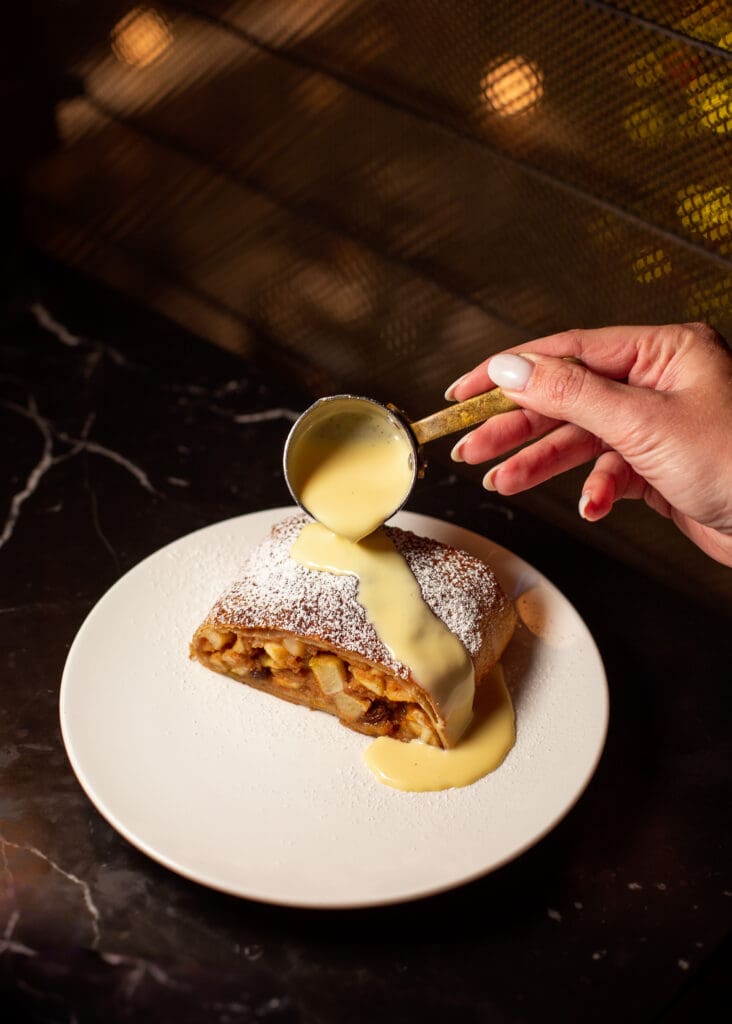 A hand pours creamy sauce from a small ladle onto a slice of apple strudel dusted with icing sugar, served on a white plate against a dark marble surface.