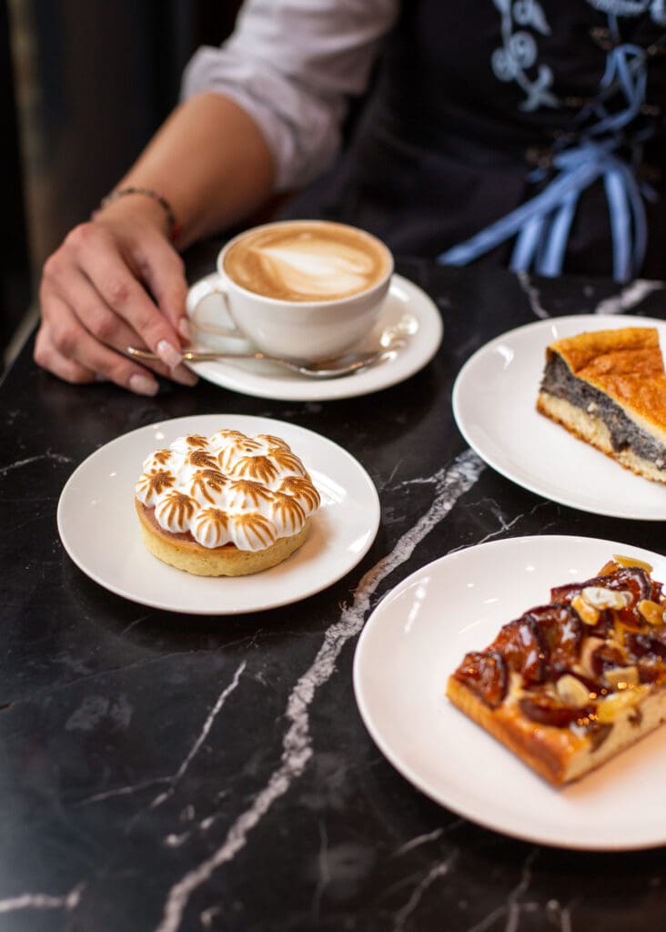 A person sitting at a black marble table with a cappuccino and three plates of desserts, including a tart topped with meringue, a dark pie slice, and a pastry with nuts and caramel.