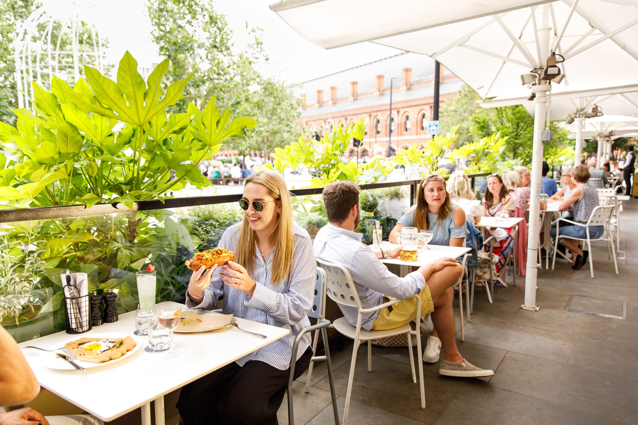 People dining outdoors at a leafy restaurant terrace on a sunny day. A woman in sunglasses eats pizza at a table; other groups are seated nearby, enjoying food and conversation under large white parasols.