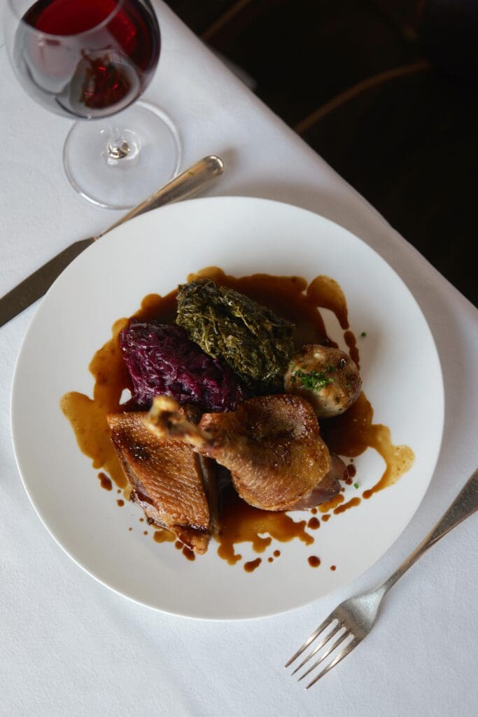A white plate with roast meat, braised red cabbage, leafy greens, and a small round dumpling, served with brown gravy, is set on a white tablecloth next to a glass of red wine, a knife, and a fork.