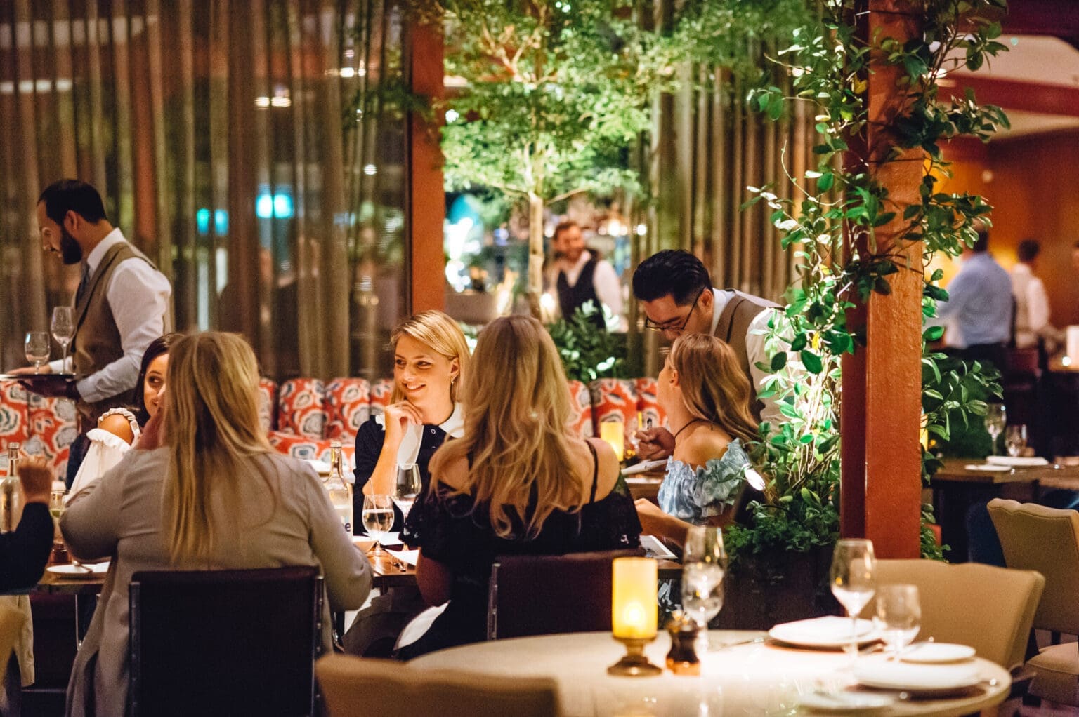 A group of people dine and chat at a lively, elegant restaurant with greenery, warm lighting, and a relaxed atmosphere; waiting staff serve food and drinks at various tables.
