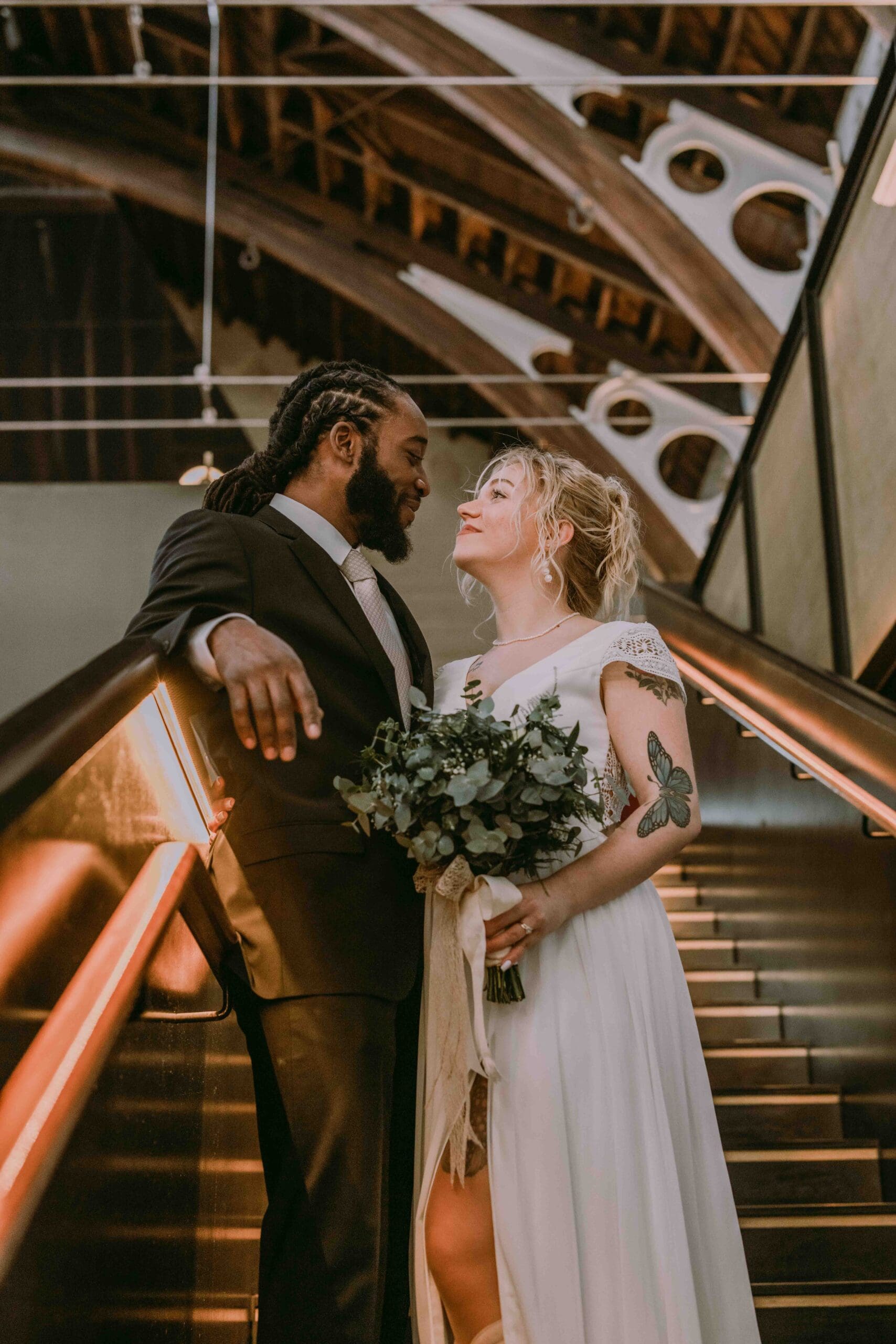 A bride and groom stand together on an escalator, gazing lovingly at each other. The bride holds a bouquet and wears a white dress, while the groom wears a suit. The background features a vaulted wooden ceiling.