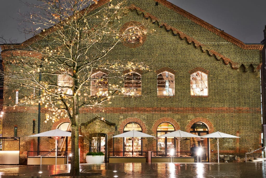 A brick building with large arched windows is illuminated at night. In front, a leafless tree with fairy lights and several white umbrellas cover outdoor seating on a wet pavement.