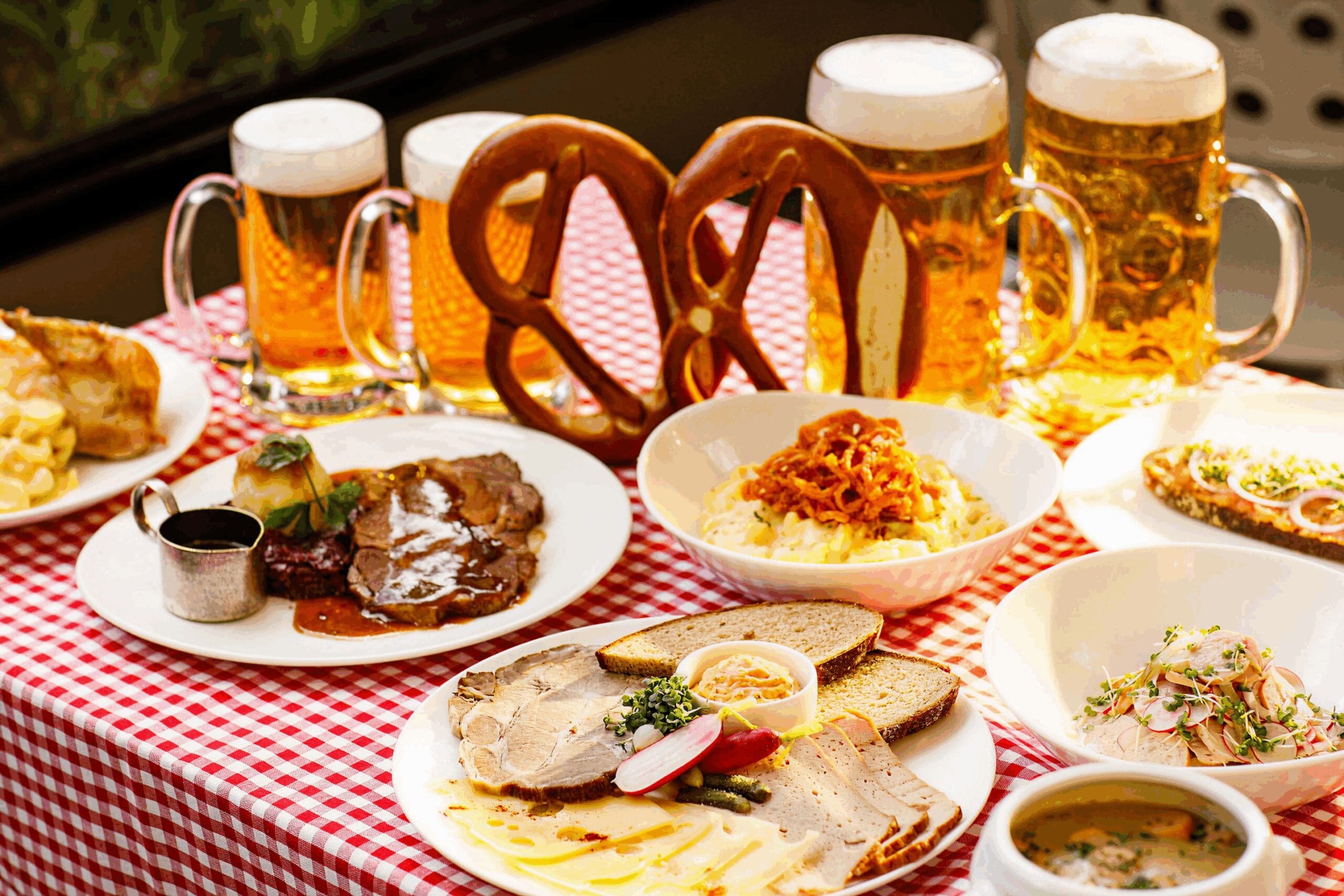 A table with German food: pretzels, roast meat with gravy, sliced meat with bread, potato salad, soup, and tankards of beer, all on a red and white checked tablecloth.