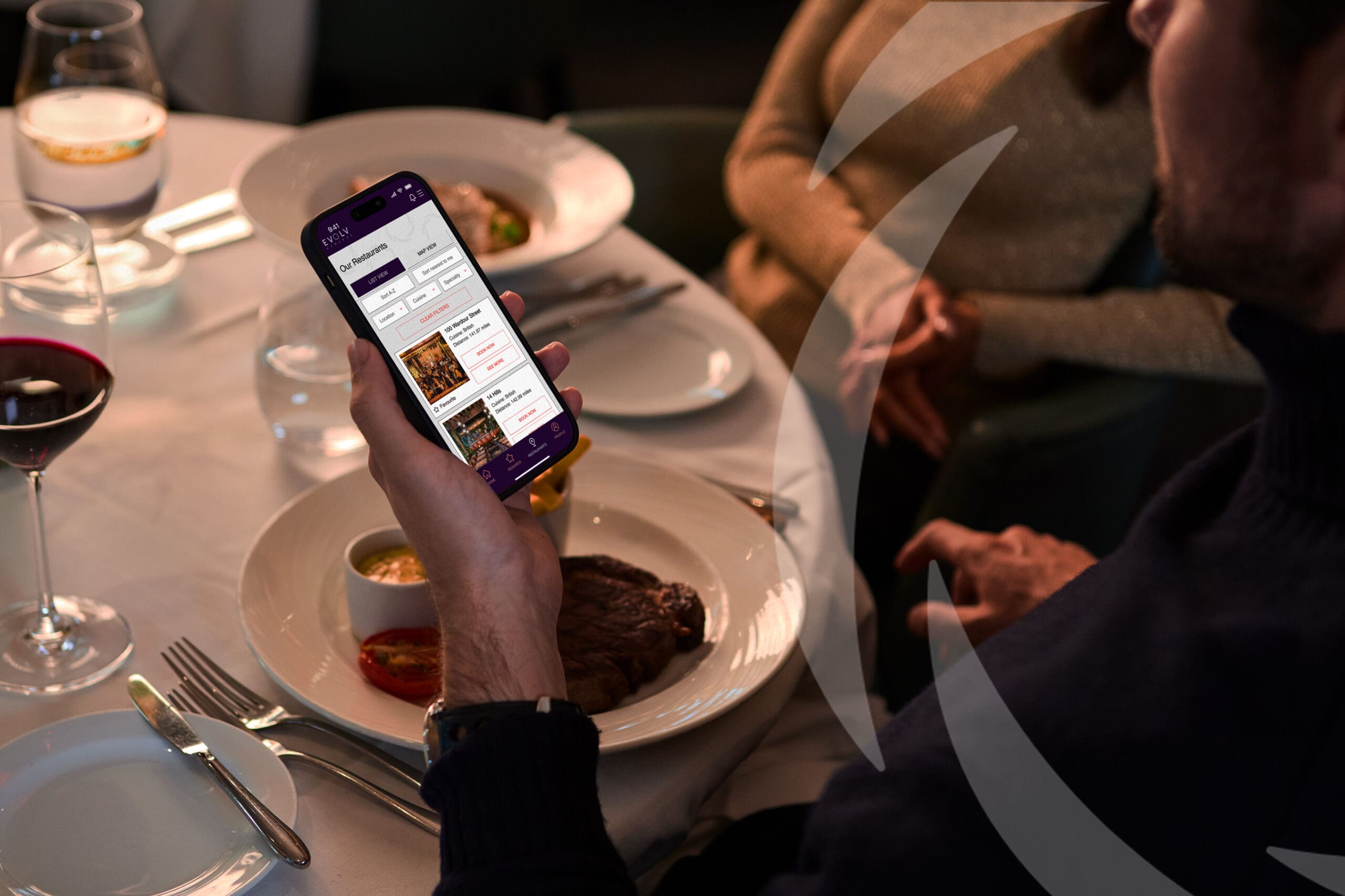 A person in a restaurant holds a mobile phone displaying a food ordering app over a table set with steak, sauce, wine, and water, whilst another person sits opposite the table.