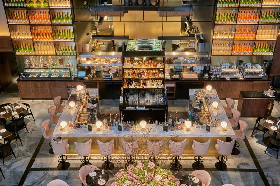 A modern restaurant interior with a large central bar surrounded by cushioned chairs, tables set for dining, decorative bottles lining the walls, and warm ambient lighting.