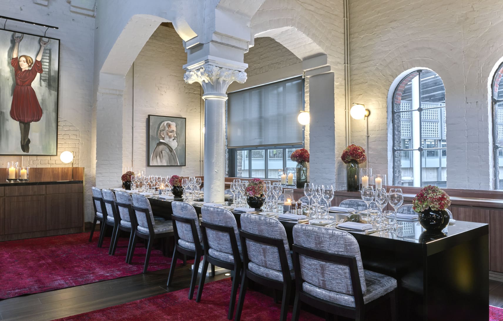 Elegant dining room with a long table set for a meal, modern chairs, red carpet, white brick walls, large arched windows, floral centrepieces, candles, and two portraits on the walls.