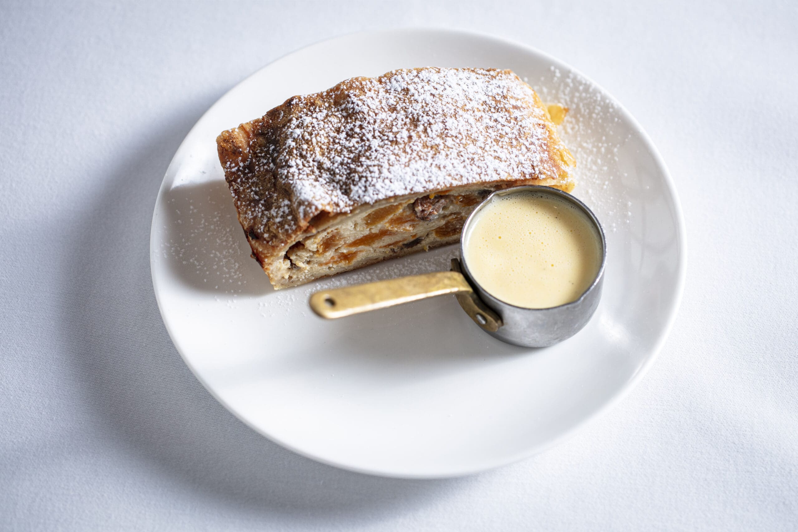 A slice of apple strudel dusted with icing sugar on a white plate, served with a small metal jug of creamy vanilla sauce.