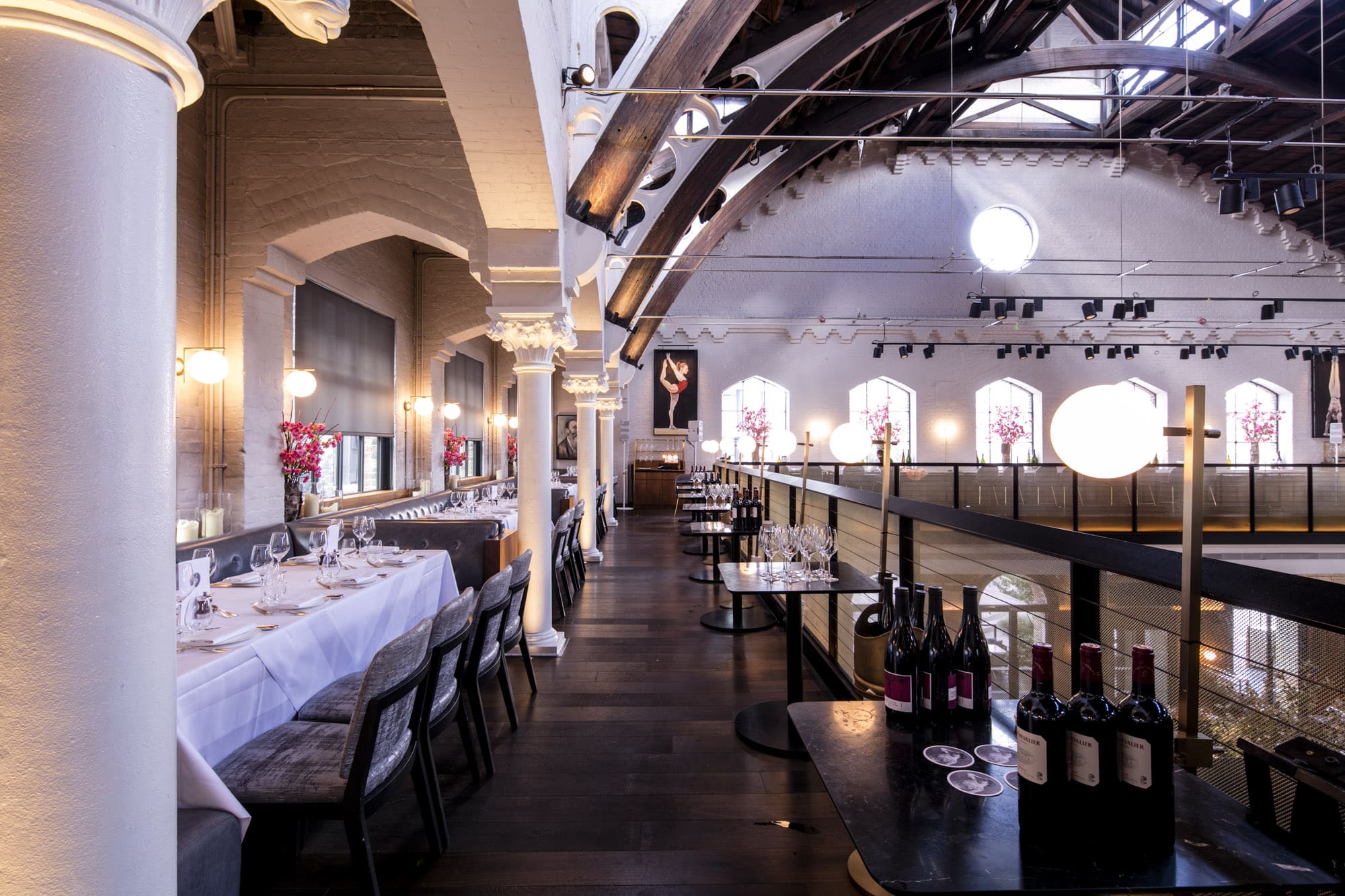 Elegant restaurant interior with high arched ceilings, exposed beams, white columns, and large windows. Tables are set with white tablecloths and wine glasses; bottles of wine are displayed on a side table.