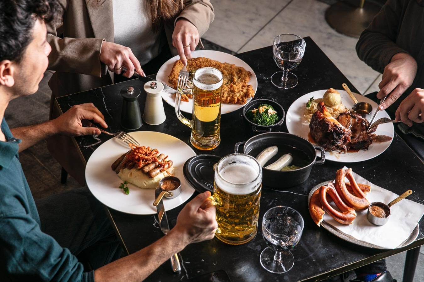 Three people share a meal at a restaurant, eating German dishes such as sausages, schnitzel, and pork knuckle. Large tankards of beer are on the table, along with glasses of water and small bowls of sauce.