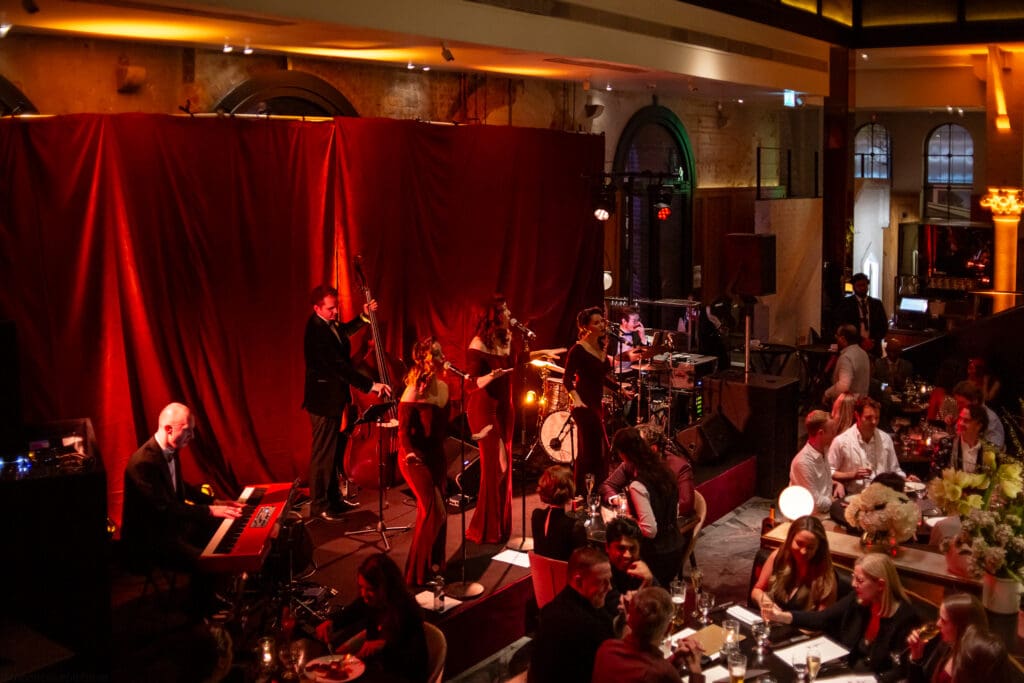 A jazz band performs on stage with red lighting in a lively restaurant, while guests sit at tables, dining and watching the show. The ambience is warm and elegant, with flowers and soft lighting.