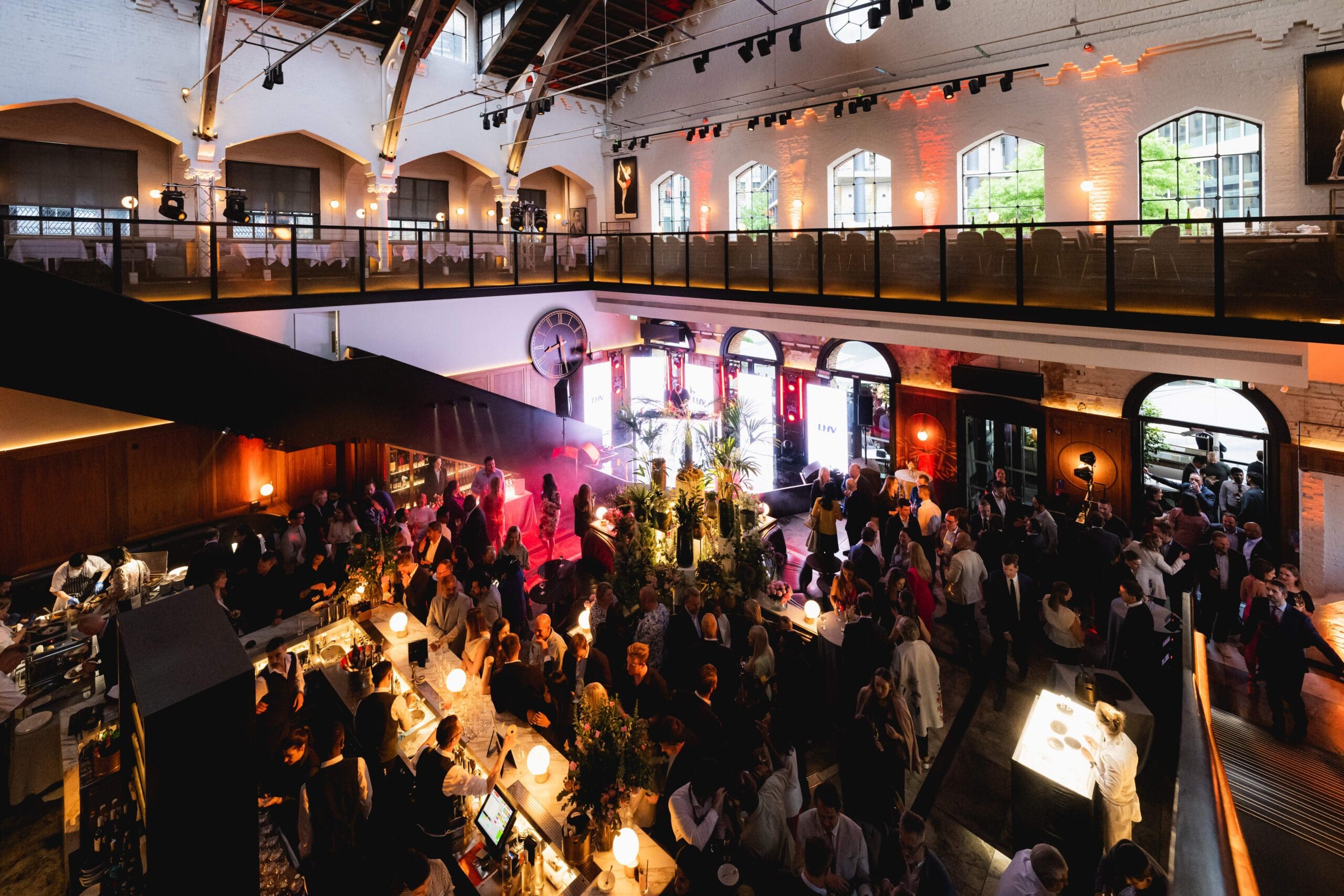 A large, stylish indoor event space filled with people socialising and mingling near a bar, with modern lighting, high ceilings, and an upper balcony area.