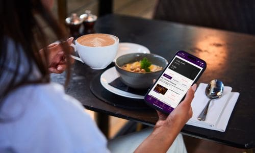 A person sits at a table holding a cup of coffee and looking at a mobile phone, which displays a ticket booking app. There is a bowl of food, a spoon, and a serviette on the table.