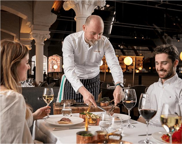 A chef in a white uniform serves food to a smiling man and woman seated at an elegant restaurant table set with wine glasses and plates. The dining room features tall columns and warm, ambient lighting.