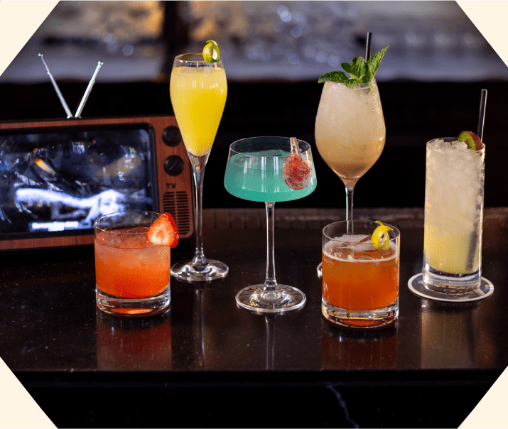 Six colourful cocktails in different glasses are displayed on a dark bar counter, with a small vintage TV in the background. Some drinks are garnished with fruit or mint, creating an elegant and lively atmosphere.