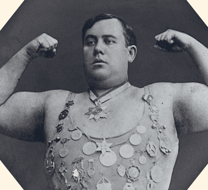 A strongman poses with arms flexed, wearing a sleeveless outfit adorned with numerous medals and a large star-shaped medallion around his neck. The image is black and white and has a vintage appearance.